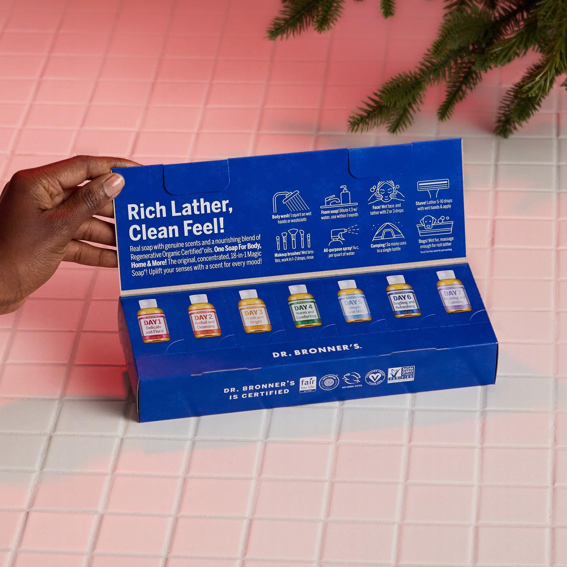 A hand holds an open blue box containing six miniature bottles of Dr. Bronners liquid castile soap varieties. The box is titled Rich Lather, Clean Feel on a tiled surface with greenery [One Size]