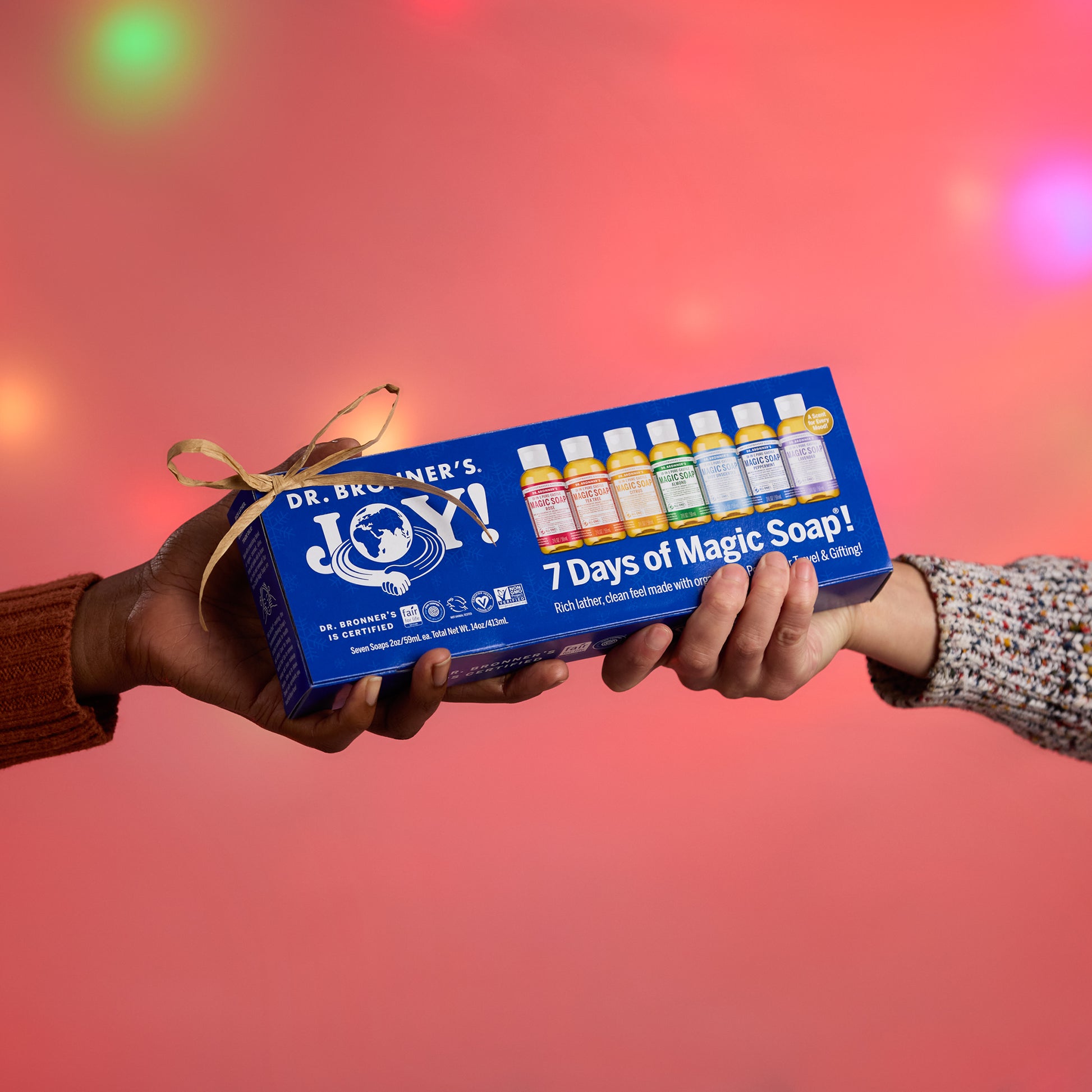 Box of Dr. Bronners castile soap bottles being handed over against a festive, blurred pink background. Dr. Bronners JOY 7 Days of Magic Soap Rich lather, clean feel made with organic oils [One Size]