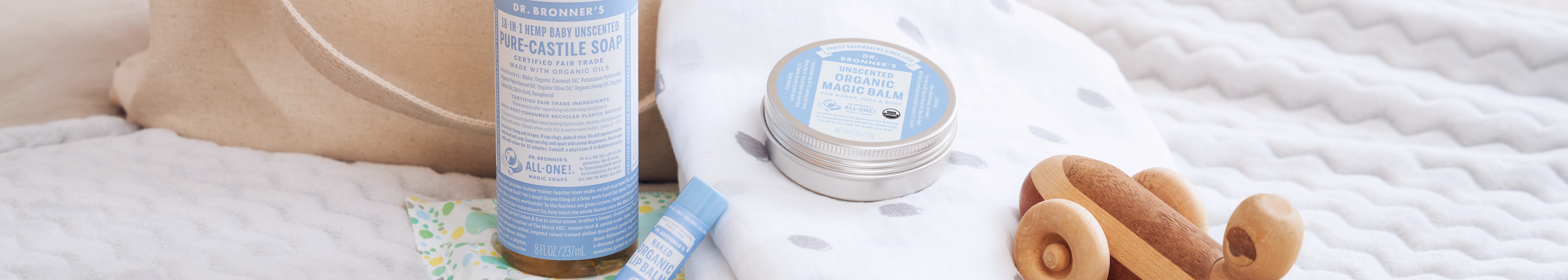 A bottle of Dr. Bronners Hemp Baby Unscented Pure Castile Soap and a tin of Unscented Organic Magic Balm are placed on a soft, white patterned fabric next to a wooden toy. DR. BRONNERS 18 IN 1 HEMP BABY UNSCENTED PURE CASTILE SOAP CERTIFIED FAIR TRADE MADE WITH ORGANIC OILS