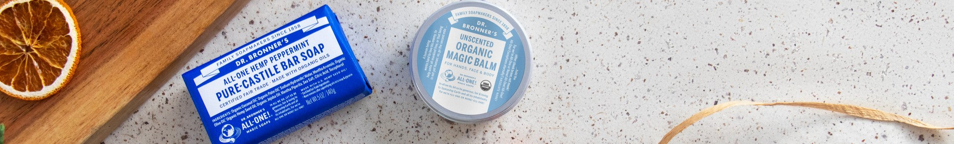 Dr. Bronners Peppermint Pure Castile Bar Soap and Unscented Organic Magic Balm sit on a speckled surface with a dried orange slice on a wooden board nearby, promoting eco friendly skincare
