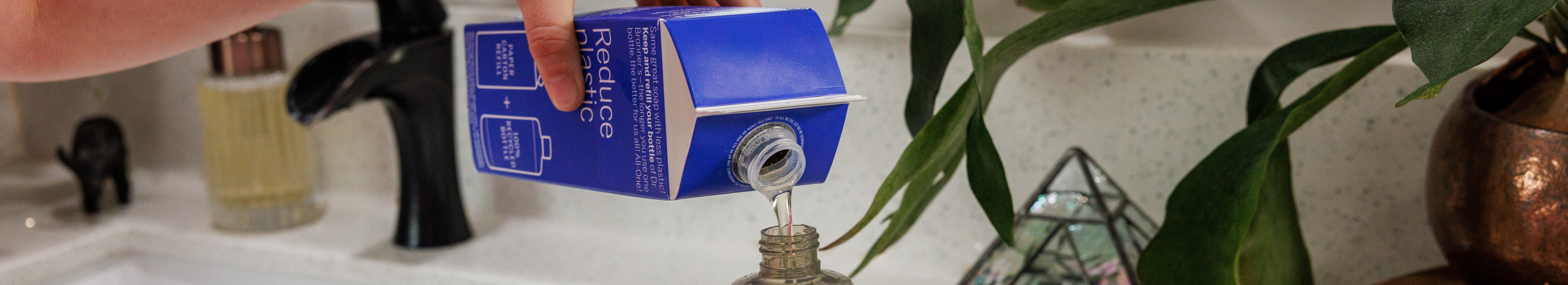 A hand pours liquid castile soap from a blue carton labeled Reduce Plastic into a glass bottle on a countertop with a plant, sink, and soap dispenser nearby