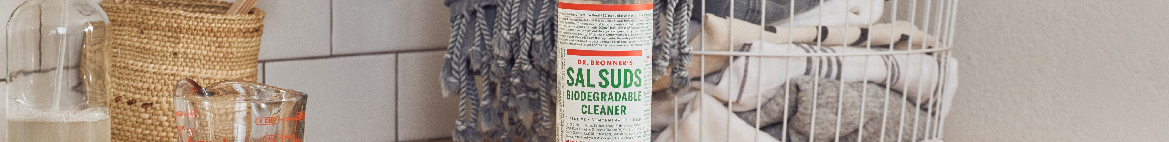 Dr. Bronners Sal Suds biodegradable cleaner is positioned on a laundry counter, next to a wicker basket and measuring cup, with a wire basket of folded towels in the background