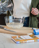 A person pours liquid into a clear bowl containing chopped ingredients on a kitchen counter. A Dr. Bronners Castile Soap box and a knife rest beside it. Vegan soap option displayed