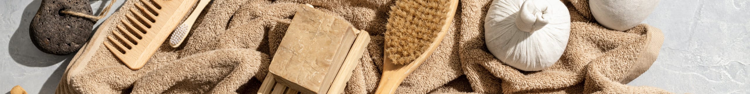 Wooden comb and pumice stone rest on a soft beige towel, accompanied by a natural bristle brush and rustic bar soap, creating a serene spa like atmosphere on a smooth surface