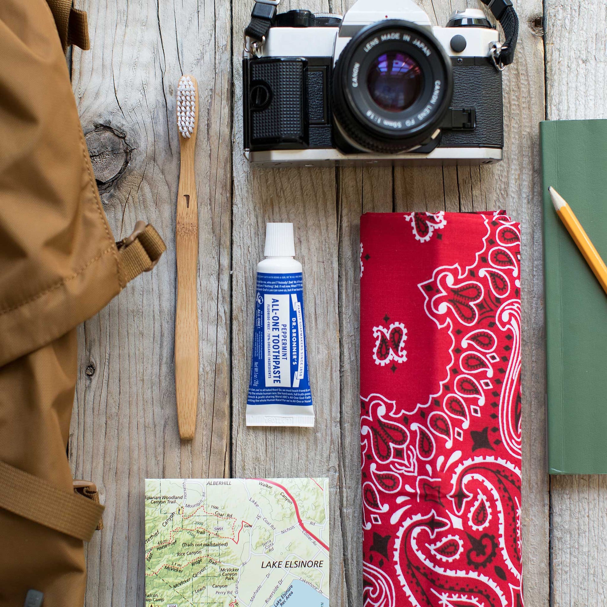 A camera, toothbrush, tube of Dr. Bronners toothpaste, map, red bandana, green notebook, and pencil are neatly arranged on a wooden surface, suggesting a travel theme. Lake Elsinore is marked on the map [5 oz]