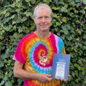 An individual in a colorful tie dye shirt holds a rectangular object with text against a lush green leafy background. The setting is outdoors, suggesting a vibrant, natural environment