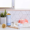 A bottle of Dr. Bronners liquid Castile soap stands beside potted herbs and a dish brush in a kitchen setting, with white tile backsplash highlighting its eco friendly design and versatile use