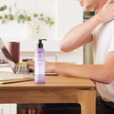 A bottle of Dr. Bronners Lavender Organic Hand Body Lotion sits on a wooden desk beside a person using a laptop, with notebooks and a plant in the background [8 oz]