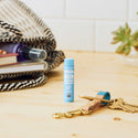 A blue tube of Dr. Bronners Naked Organic Lip Balm stands on a wooden table, surrounded by keys, books, and a woven bag, with a potted plant in the background [.15 oz]