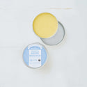 A round tin container with its lid off reveals a yellow balm inside. The label reads, Dr. Bronners Magic Balm, Baby Unscented. Its set on a light surface  [2 oz]