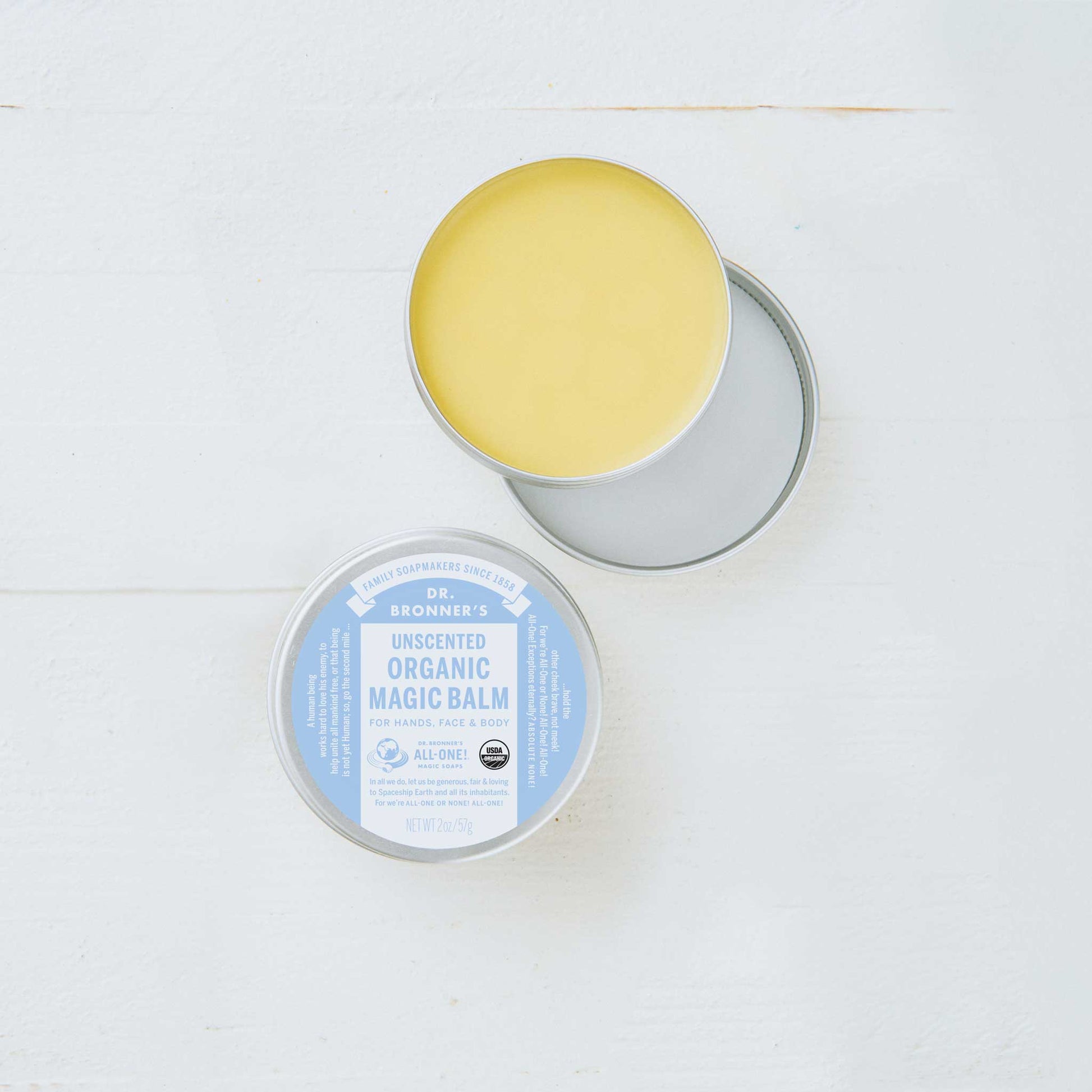 A round tin container with its lid off reveals a yellow balm inside. The label reads, Dr. Bronners Magic Balm, Baby Unscented. Its set on a light surface  [2 oz]