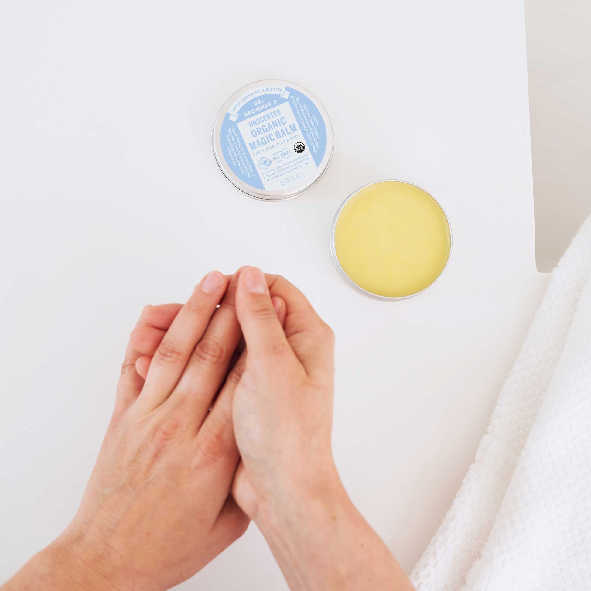 Hands apply balm from an open tin of Dr. Bronners Organic Magic Balm with a blue label on a white surface, next to a textured cloth  [2 oz]