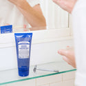 A tube of Dr. Bronners Peppermint Organic Shaving Soap sits on a bathroom glass shelf, accompanied by a razor. A person is using the soap, reflected in the mirror [7 oz]