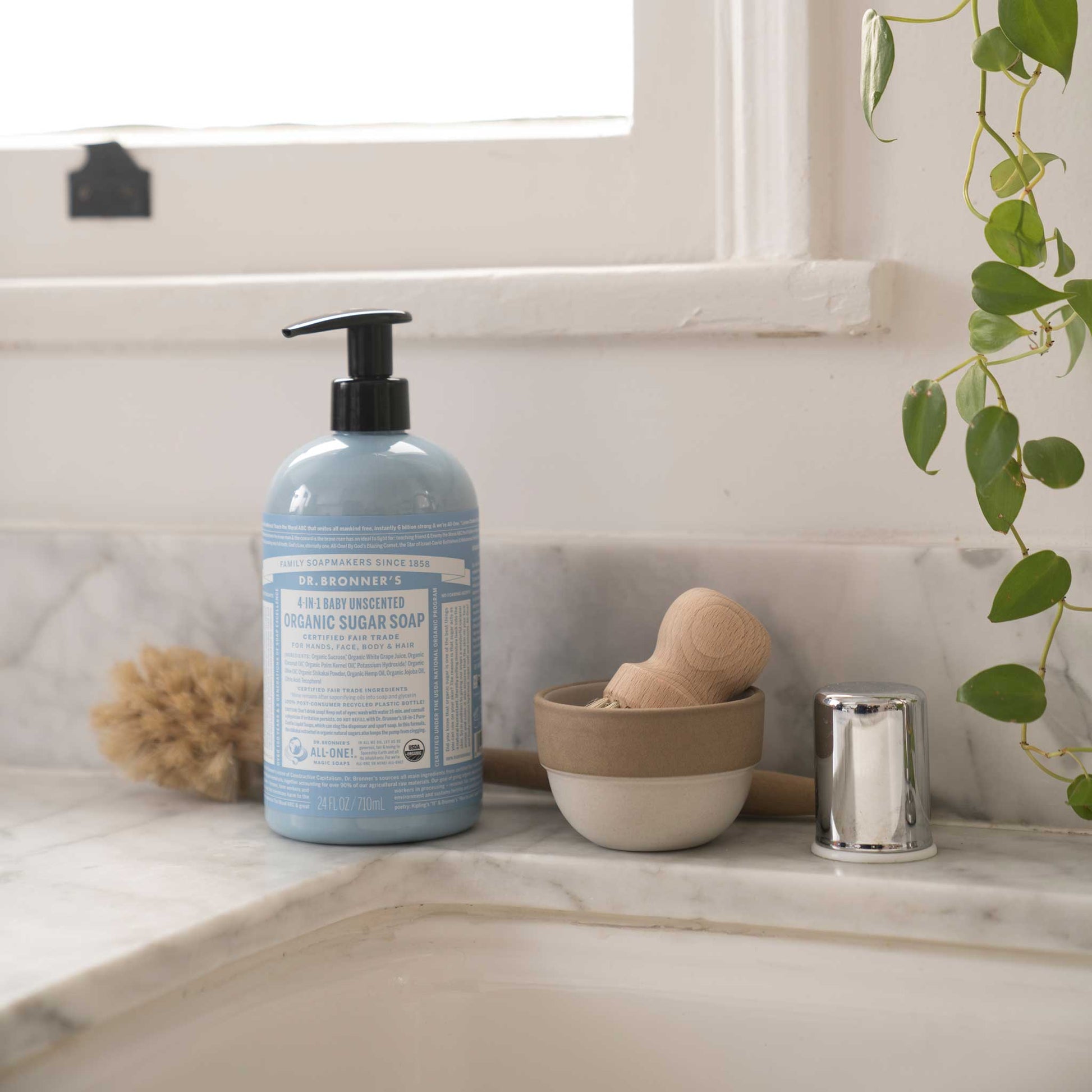 A bottle of Dr. Bronners liquid soap stands on a marble countertop beside grooming tools. The sunlit window and green plant enhance an eco friendly bathroom ambiance. Text on the bottle includes product details [24 oz]