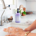Hands are being washed under a running tap with Dr. Bronners 4 in 1 Lavender Organic Sugar Soap on a marble counter. Nearby, a plant, bowl, and towel enhance the bathroom decor [24 oz]