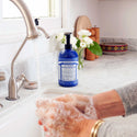 A person washes hands with foamy soap in a kitchen sink. Dr. Bronners 4 in 1 Peppermint Organic Sugar Soap sits on the counter beside flowers and kitchen items [24 oz]