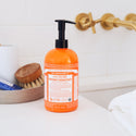 Dr. Bronners 4 in 1 Tea Tree Organic Sugar Soap bottle sits on a bathroom counter. Gold faucets and a tray with grooming items provide a minimalistic, serene setting. 24 fl oz710 mL  [24 oz]