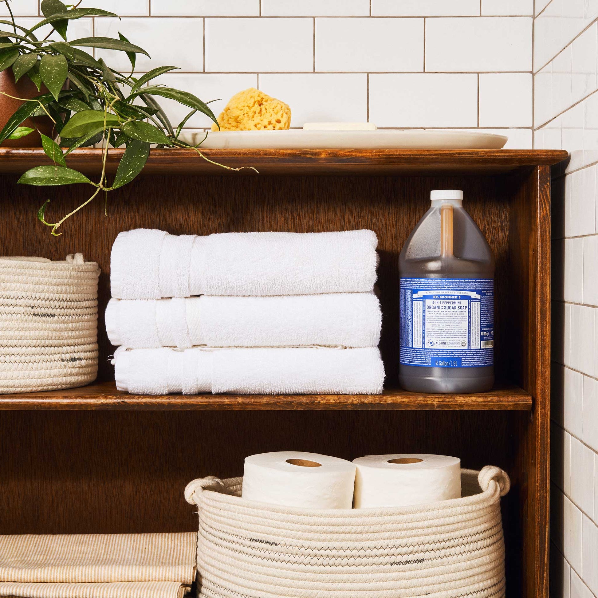 A bottle of Dr. Bronners liquid castile soap sits on a wooden bathroom shelf beside neatly folded white towels. Nearby, wicker baskets hold toilet paper and a potted plant enhances the natural, organized setting [1/2 Gallon]