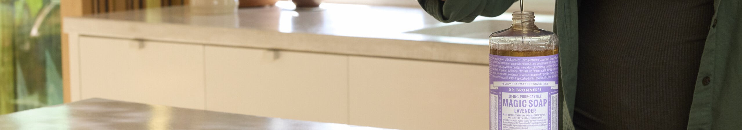 A person fills a dispenser with liquid from a large bottle of Dr. Bronners 18 in 1 Pure Castile Magic Soap Lavender in a modern kitchen setting. Text on the bottle reads Dr. Bronners Family Soapmakers Since 1858, 18 in 1 Pure Castile Magic Soap Lavender, Made With Organic Oils followed by more detailed information