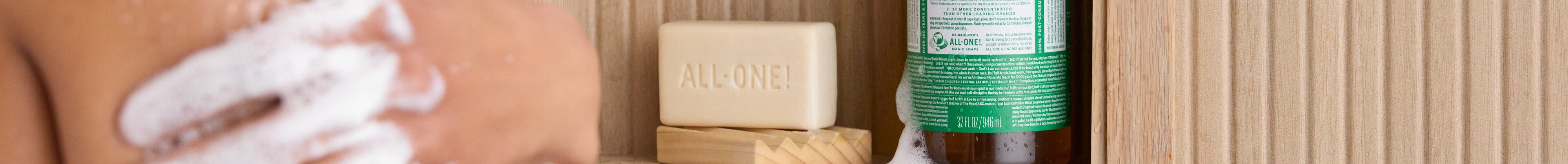 A person is lathering their body with foam. Nearby, a bar of soap labeled ALL ONE and a bottle of liquid soap with a green label are placed on a wooden surface