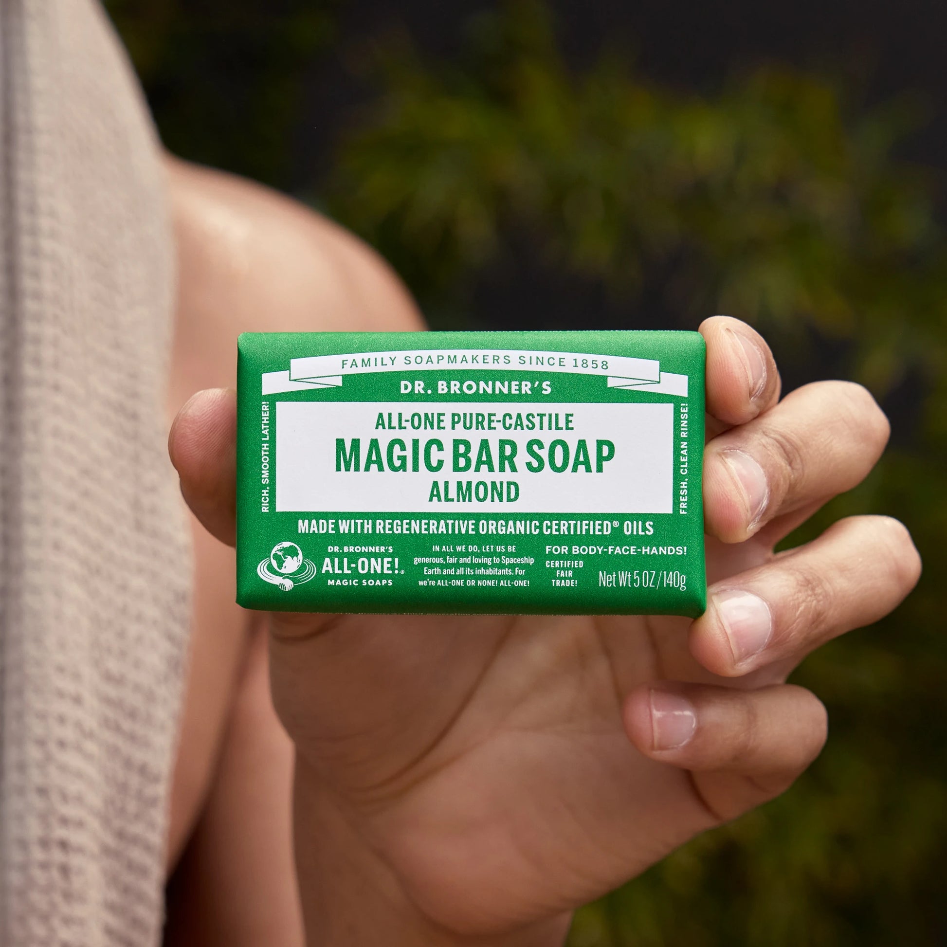 A hand holds a green package labeled Dr. Bronners All One Pure Castile Magic Bar Soap Almond, highlighting its regenerative organic certified oils, suitable for body, face, and hands. The package states Family Soapmakers Since 1858. Rich. Smooth. Lather Fresh. Clean. Rinse Certified Fair Trade. Net Wt 5.0 oz140 g [5 oz]