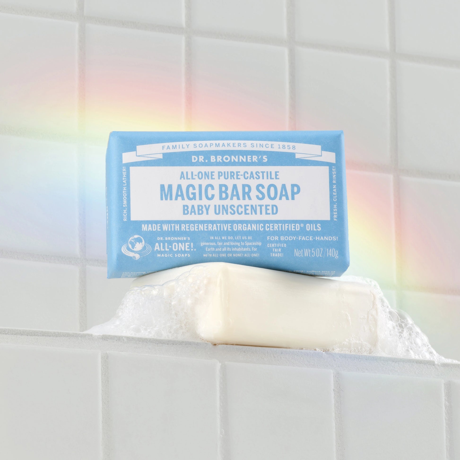 A Dr. Bronners Baby Unscented Magic Bar Soap rests on a wet, tiled surface with visible suds, accompanied by a rainbow reflection. Family Soapmakers Since 1858. Dr. Bronners All One Pure Castile Magic Bar Soap Baby Unscented. Made with regenerative organic certified oils. For body, face, hair Made in USA. NET WT 5 OZ 140g [5 oz]