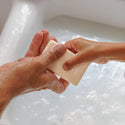 Hands hold a bar of soap over a sudsy sink, exchanging it in a gentle motion. The scene conveys cleanliness and trust, ideal for organic castile soap promoting sustainable, vegan body care solutions [5 oz]