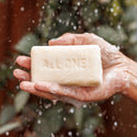 A persons hands hold a frothy bar of soap with ALL ONE engraved, amidst water droplets. The background shows green foliage, suggesting an outdoor setting [5 oz]