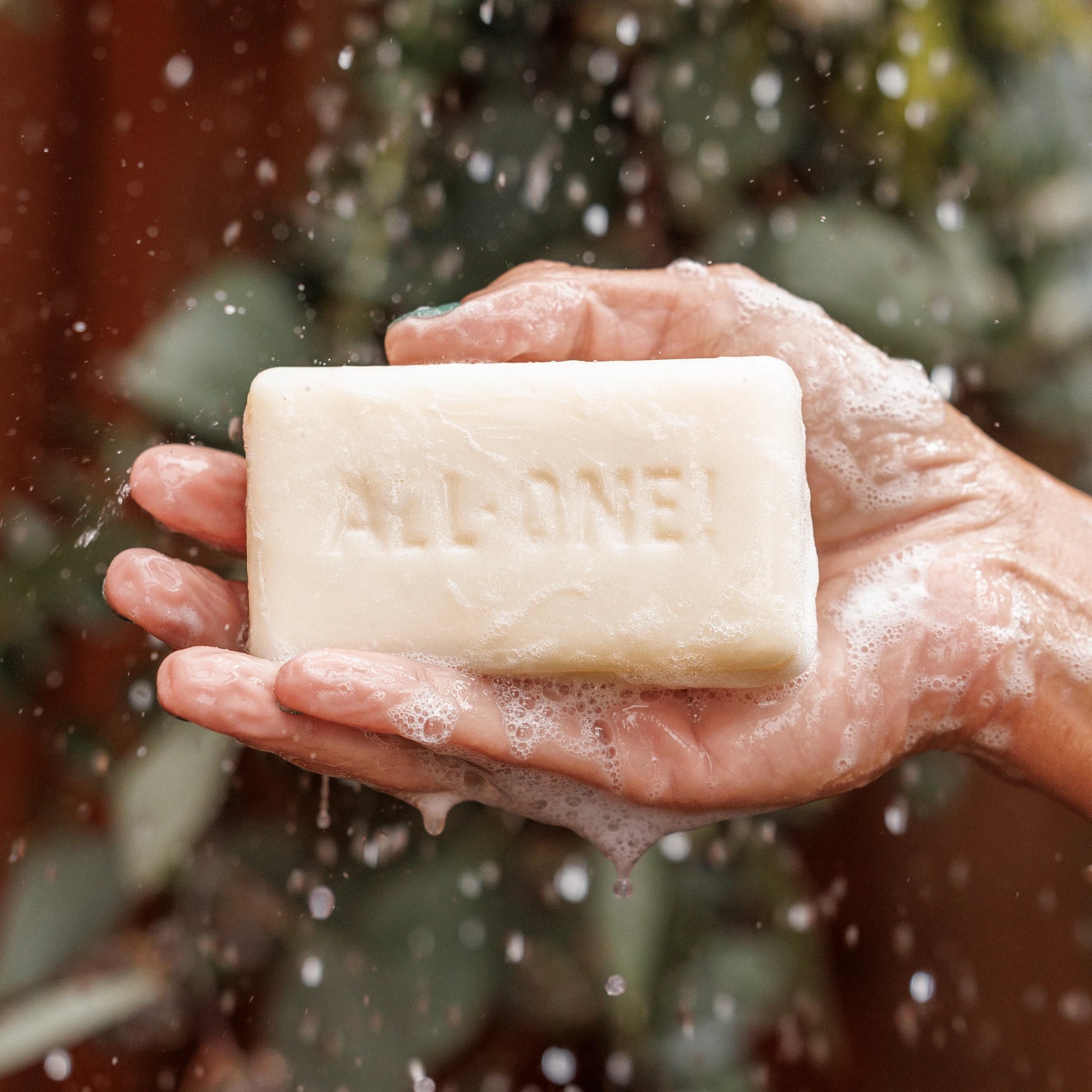 A persons hands hold a frothy bar of soap with ALL ONE engraved, amidst water droplets. The background shows green foliage, suggesting an outdoor setting [5 oz]