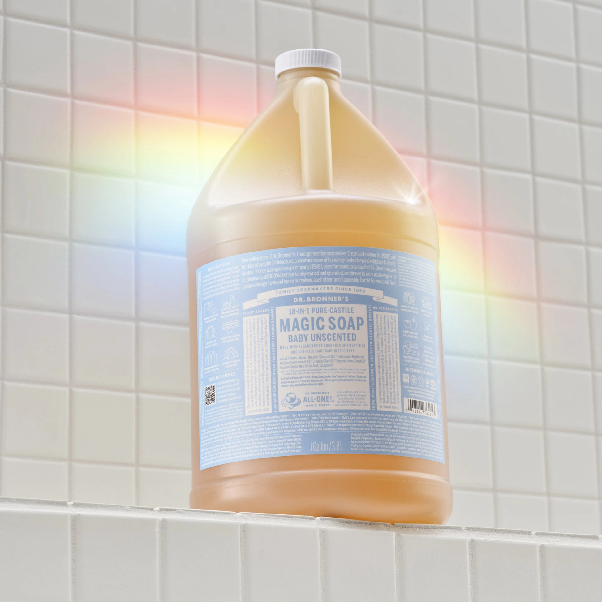 A large bottle of Dr. Bronners liquid castile soap rests on a tiled bathroom shelf, illuminated by a colorful rainbow reflection. The label includes text, but specific details are not legible [1 Gallon]