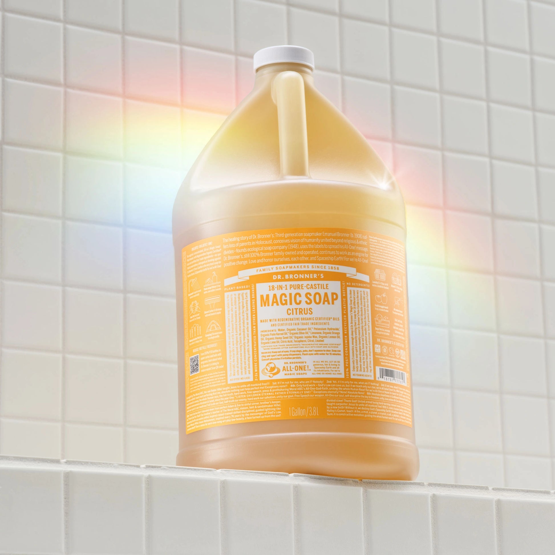A large bottle of Dr. Bronners 18 in 1 Citrus Magic Soap sits on a tiled surface, illuminated by a rainbow light. The label details various uses, emphasizing eco friendly and organic ingredients, and the bottle capacity reads 1 Gallon 3.8 L [1 Gallon]