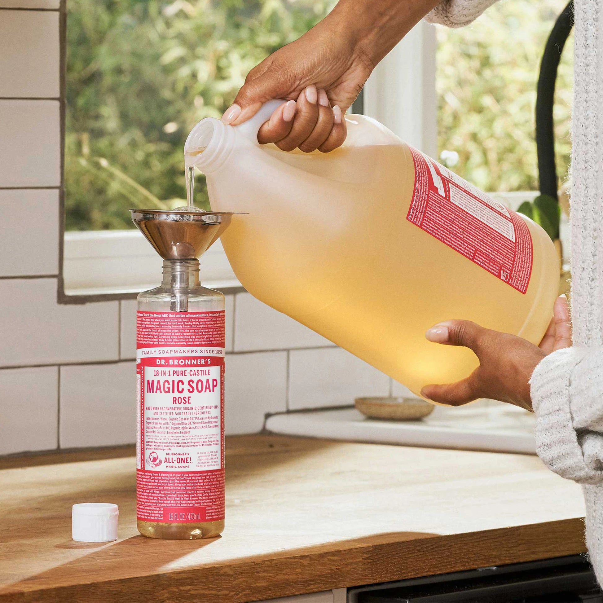 A person refills a Dr. Bronners bottle labeled 18 IN 1 Hemp Rose Pure Castile Soap from a large jug using a funnel. The setting is a kitchen with visible sink and counter [16 oz, 1 Gallon]
