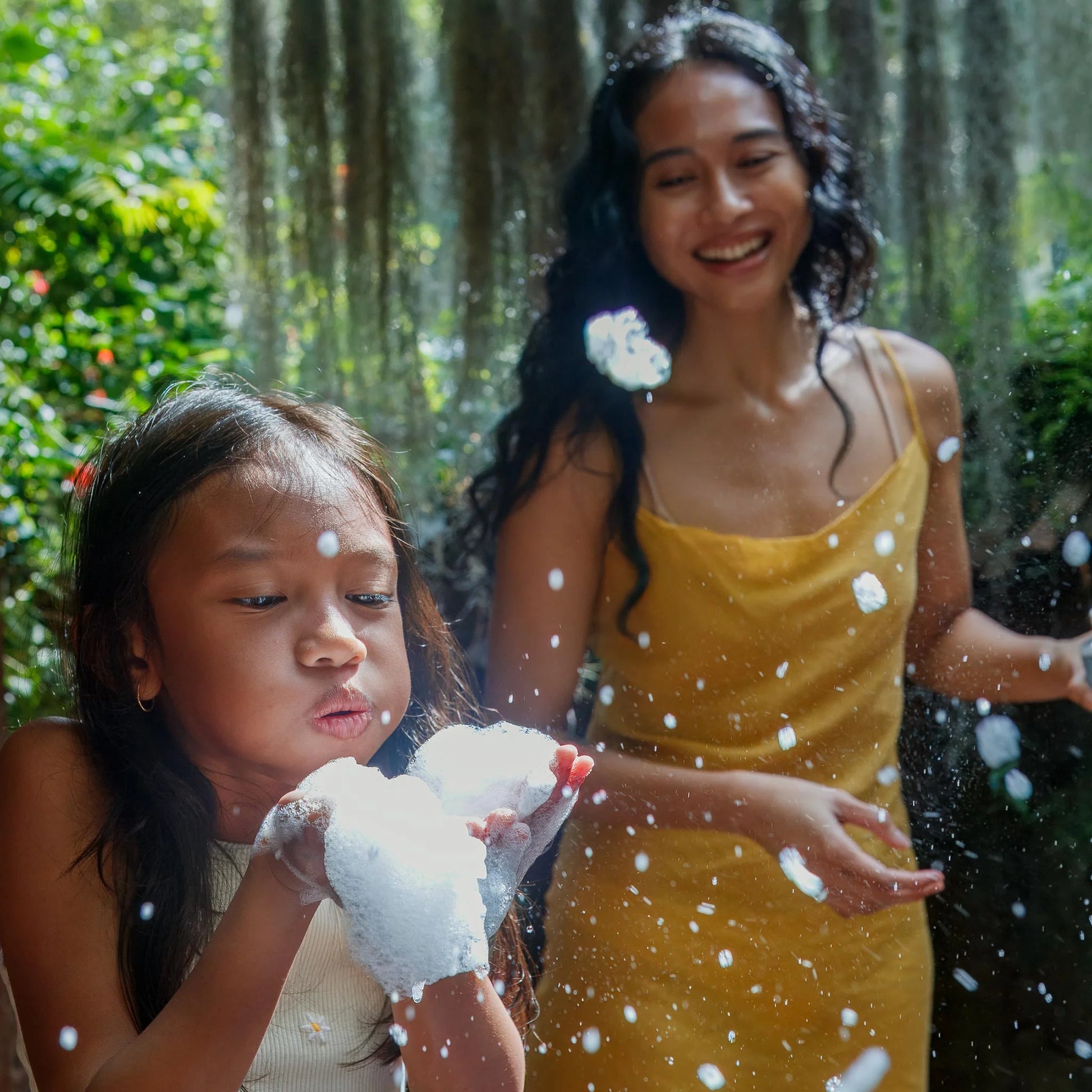 A child blows soap bubbles from her hands, creating foam while a woman in a yellow dress smiles nearby in a lush, sunlit garden [2 oz, 3.4 oz, 4 oz, 8 oz, 16 oz, 32 oz, 1 Gallon]