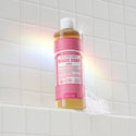 A bottle of Dr. Bronners Rose Castile Soap stands upright on a white tiled surface. A subtle rainbow reflection graces the scene, adding a vibrant touch to the minimalist bathroom setting  [16 oz]