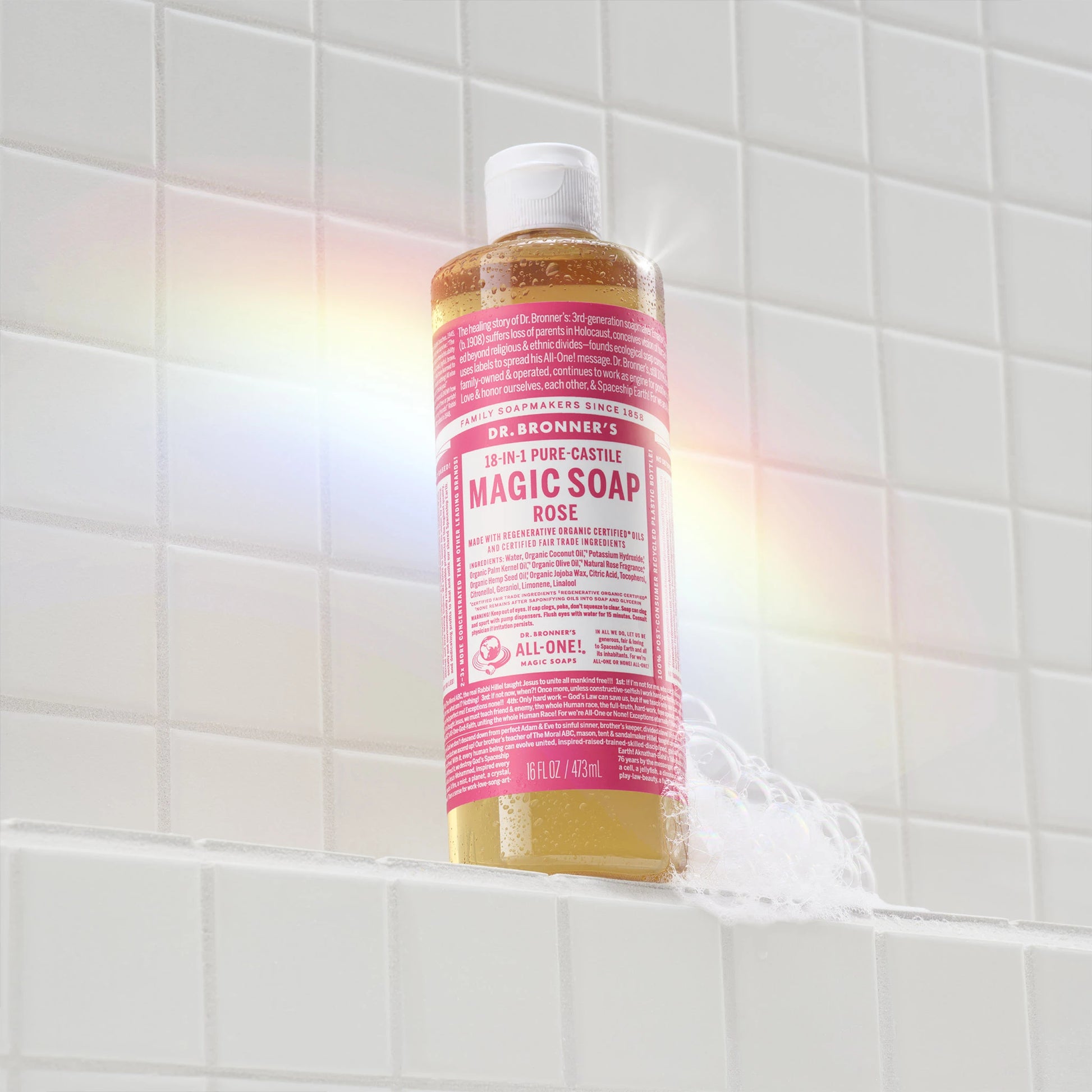 A bottle of Dr. Bronners Rose Castile Soap stands upright on a white tiled surface. A subtle rainbow reflection graces the scene, adding a vibrant touch to the minimalist bathroom setting  [16 oz]