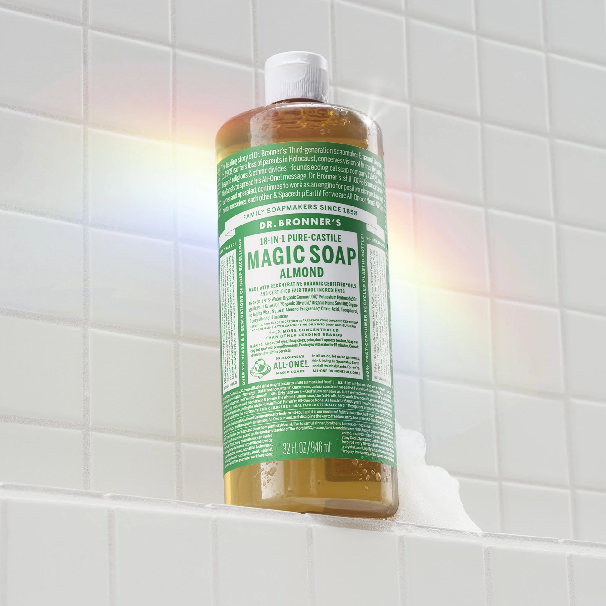 A bottle of Dr. Bronners Pure Castile Soap stands upright on a tiled bathroom shelf, with a rainbow light effect on the tiles. Text reads ALL ONE MAGIC SOAPS DR. BRONNERS PURE CASTILE SOAP DR. BRONNERS ALL ONE FAITH SERVICE SIGHT LOVE ALL ONE OR NONE LET THE ALL ONE VISION LIGHT UP STRONG BRIGHT ALL ONE [32 oz]