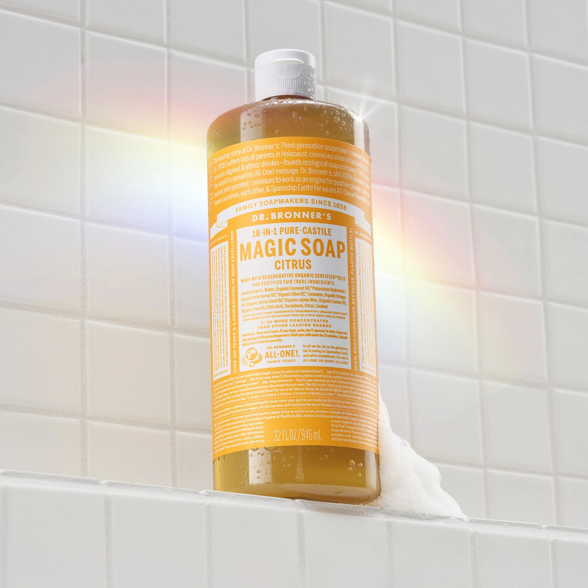 A bottle of Dr. Bronners 18 in 1 Pure Castile Magic Soap Citrus stands on a tiled surface with soap bubbles forming around its base, illuminated by soft rainbow light [32 oz]