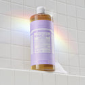 A bottle of Dr. Bronners lavender liquid castile soap stands on a white tiled shelf, illuminated by a soft rainbow reflection. Text on the bottle DR. BRONNERS 18 in 1 Pure Castile Soap [32 oz]