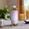 A bottle of Dr. Bronners 18 in 1 Pure Castile Magic Soap in lavender scent stands on a rustic board in a cozy room with candles and greenery, featuring 32 fl oz capacity [32 oz]