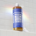 A blue bottle of Dr. Bronners Peppermint Pure Castile Liquid Soap stands on a white tiled surface, surrounded by a subtle rainbow reflection. Text on the bottle Dr. Bronners All One Magic Soap Peppermint [32 oz]