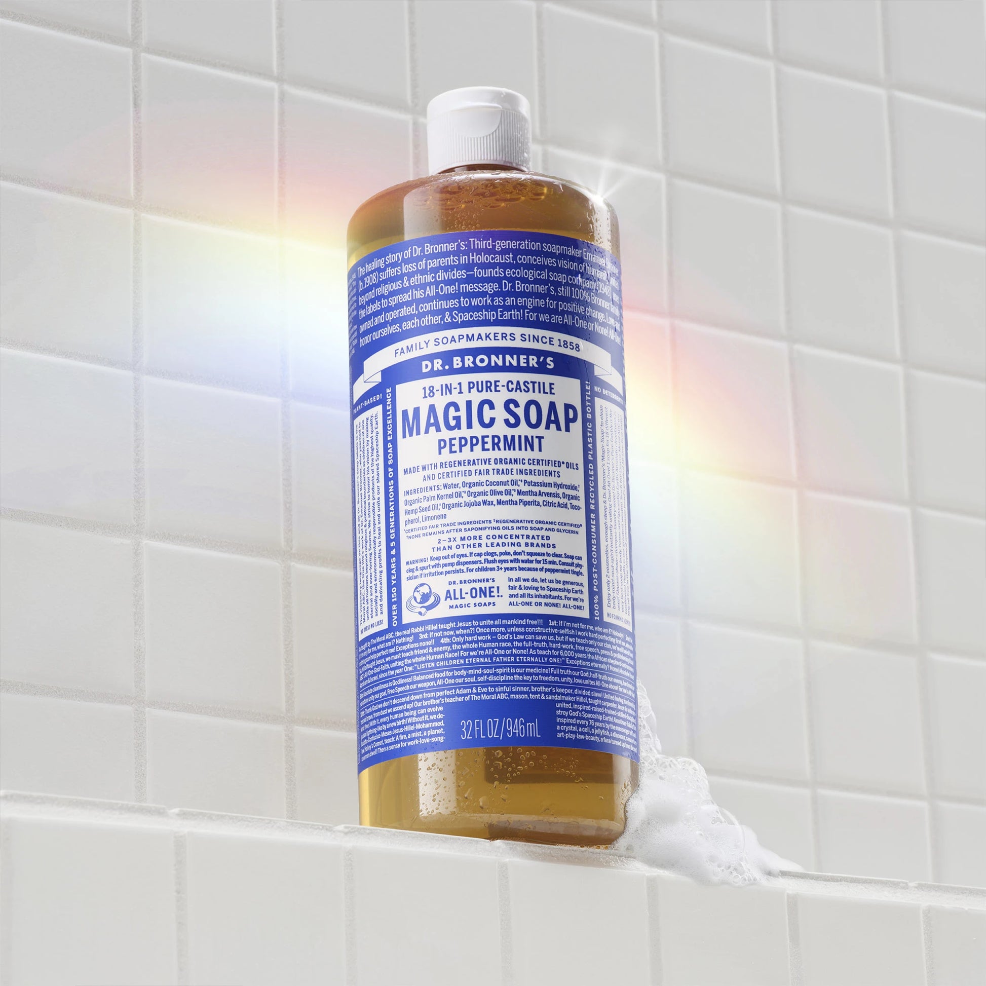 A blue bottle of Dr. Bronners Peppermint Pure Castile Liquid Soap stands on a white tiled surface, surrounded by a subtle rainbow reflection. Text on the bottle Dr. Bronners All One Magic Soap Peppermint [32 oz]