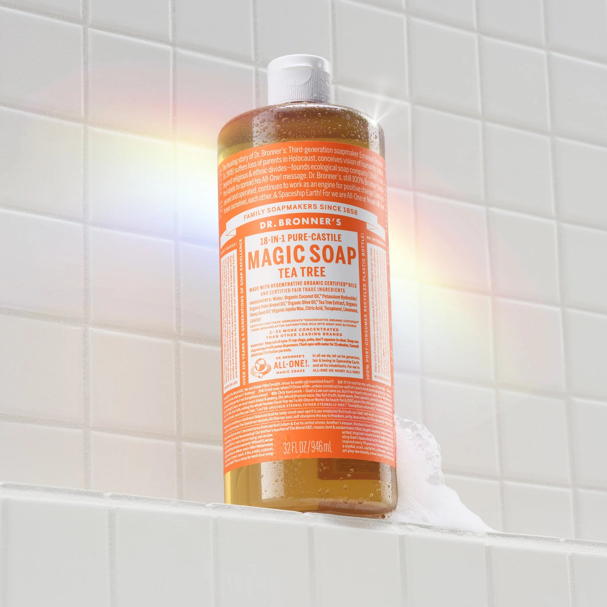 A bottle of Dr. Bronners 18 in 1 Pure Castile Magic Soap in tea tree scent rests on a soapy white tiled bathroom shelf, capturing a subtle rainbow reflection. Dr. Bronners 18 in 1 Pure Castile Magic Soap Tea Tree [32 oz]