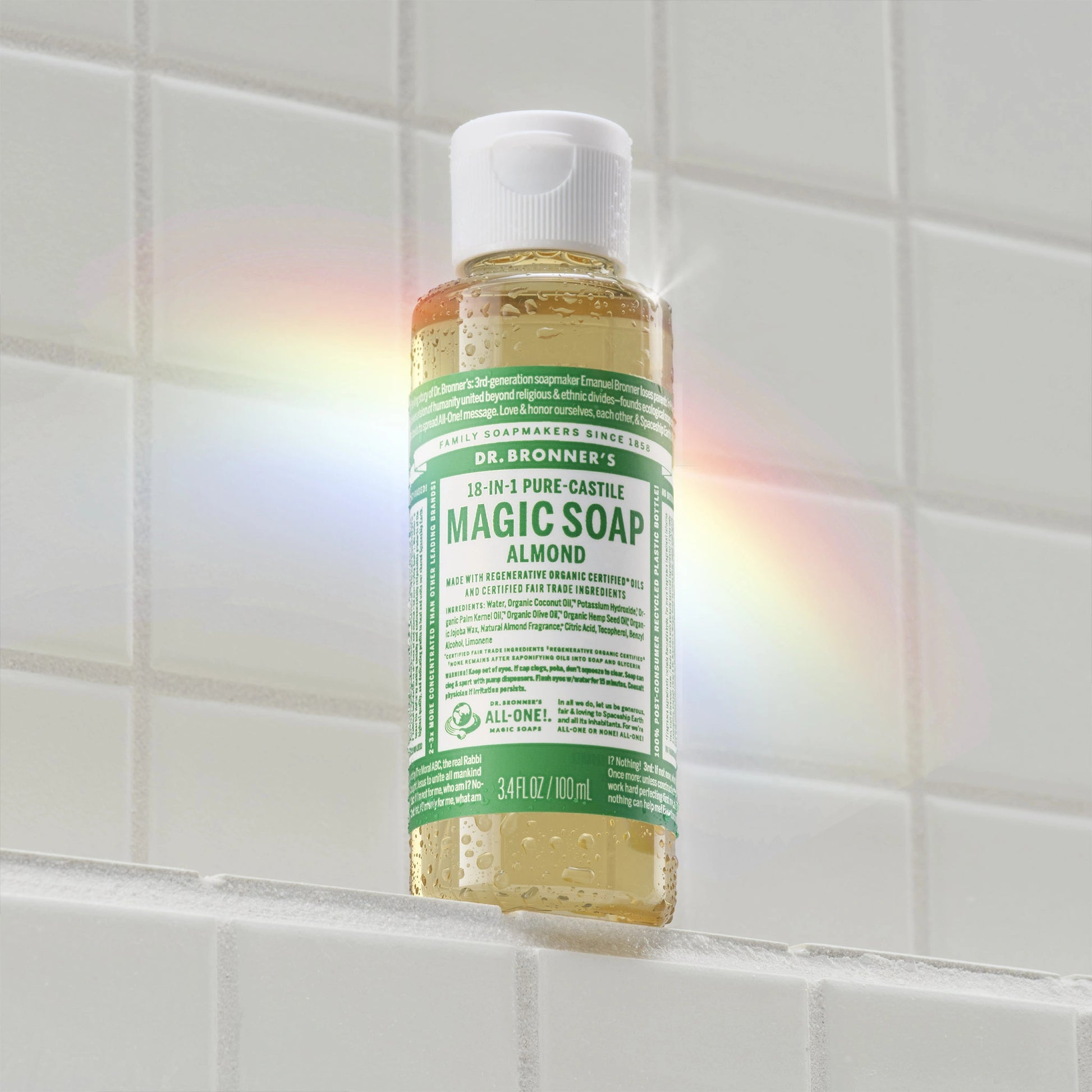 A bottle of Dr. Bronners Magic Soap with a green label, floating against white tiled background, casts a rainbow reflection. Text on bottle Magic Soap, Pure Castile Soap, 18 in 1 Hemp [3.4 oz]