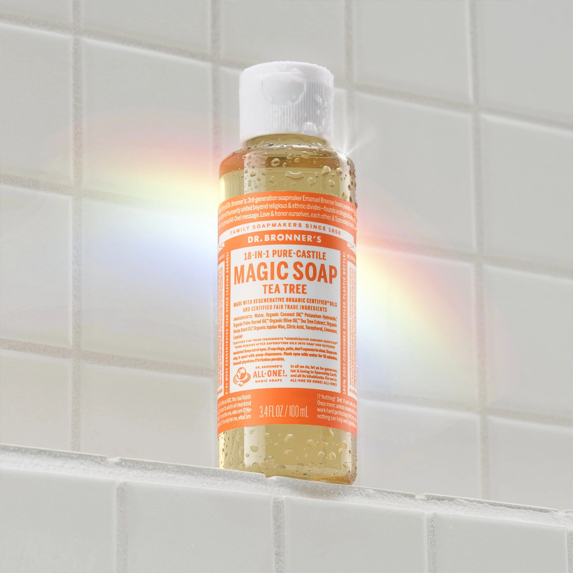 A bottle of Dr. Bronners 18 in 1 Pure Castile Tea Tree Magic Soap stands on a tiled surface, with a rainbow reflection casting on the white wall. DR. BRONNERS 18 IN 1 PURE CASTILE MAGIC SOAP TEA TREE and 3 FL OZ 89 mL [3.4 oz]