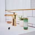 A small bottle of Dr. Bronners liquid soap stands on a white sink beside a gold faucet, accompanied by a potted plant. The bathroom features white tile and natural lighting [4 oz]