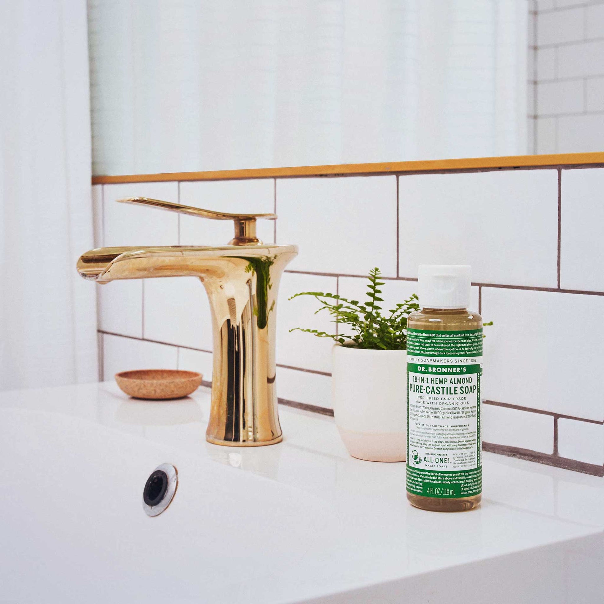 A small bottle of Dr. Bronners liquid soap stands on a white sink beside a gold faucet, accompanied by a potted plant. The bathroom features white tile and natural lighting [4 oz]
