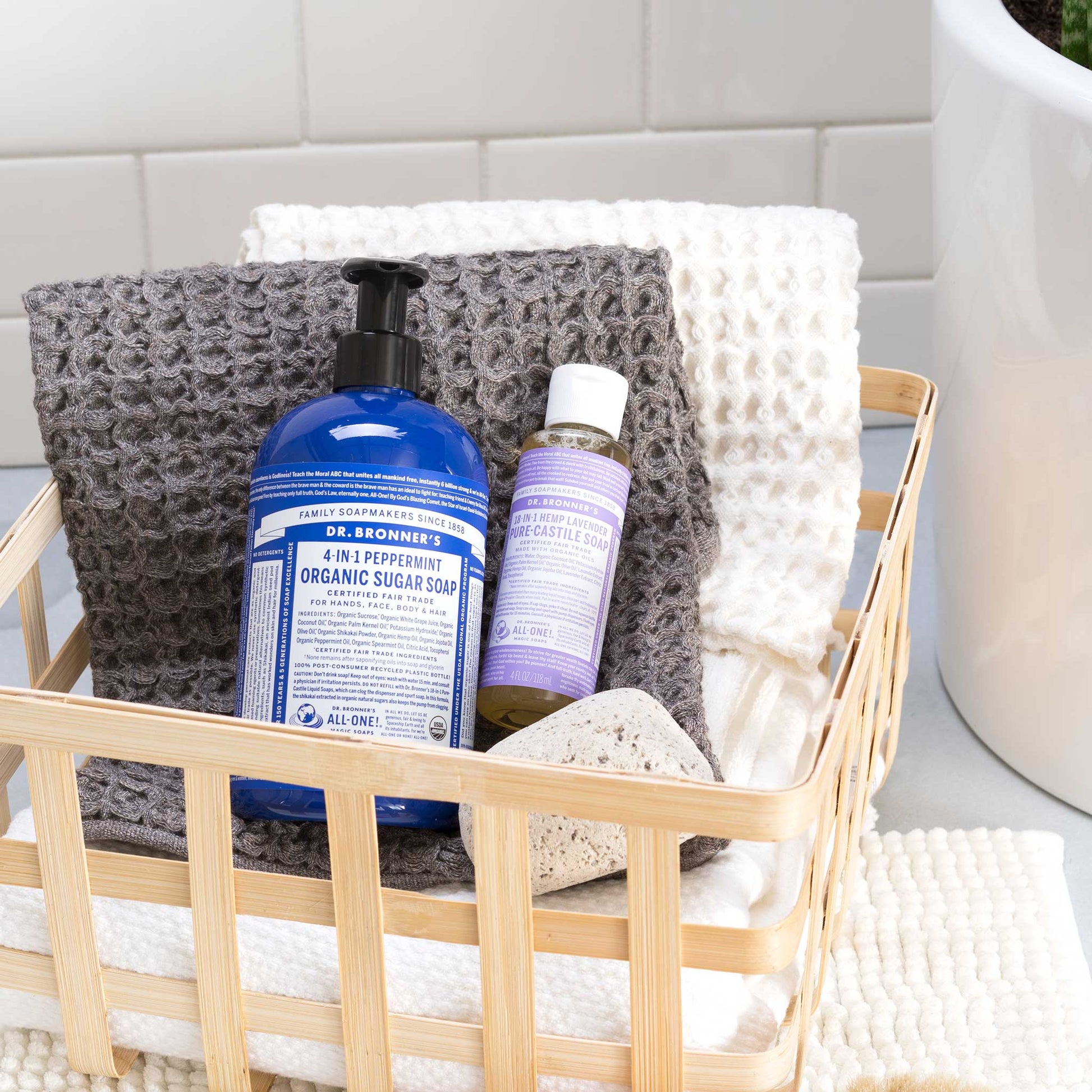 A bamboo basket holds Dr. Bronners 4 in 1 Peppermint Organic Sugar Soap and 18 in 1 Hemp Lavender Pure Castile Soap, atop textured towels beside a white tiled wall. DR. BRONNERS 4 IN 1 PEPPERMINT ORGANIC SUGAR SOAP DR. BRONNERS 18 IN 1 HEMP LAVENDER PURE CASTILE SOAP [4 oz]
