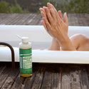A bottle of Dr Bronners liquid castile soap sits on a wooden deck beside a bathtub. A person in the tub clasps their hands, set against a natural, outdoor backdrop [8 oz]