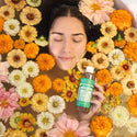 A person lies in a flower filled bath holding a bottle of Dr. Bronners Magic Soap. The colorful flowers create a serene and natural setting for relaxation and skincare. Text on the bottle reads Dr. Bronners 18 in 1 Hemp Peppermint Pure Castile Soap [8 oz]