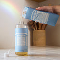 A bottle of Dr. Bronners baby unscented castile soap being refilled on a kitchen counter, with a rainbow reflection on the wall. A container with utensils is in the background [32 oz]